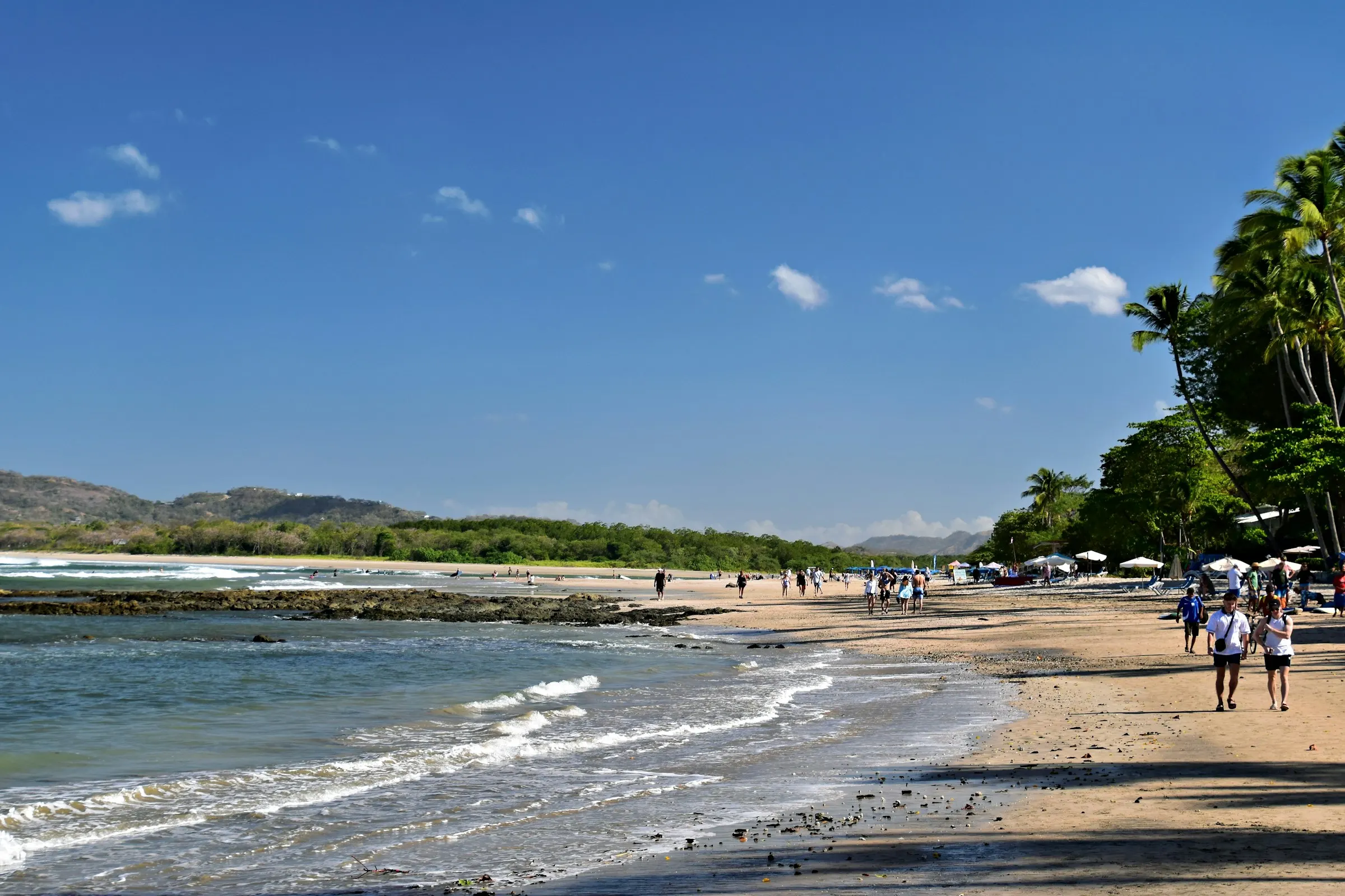 Wide sunlit beach at Tamarindo with people walking the shoreline and palms along the treeline